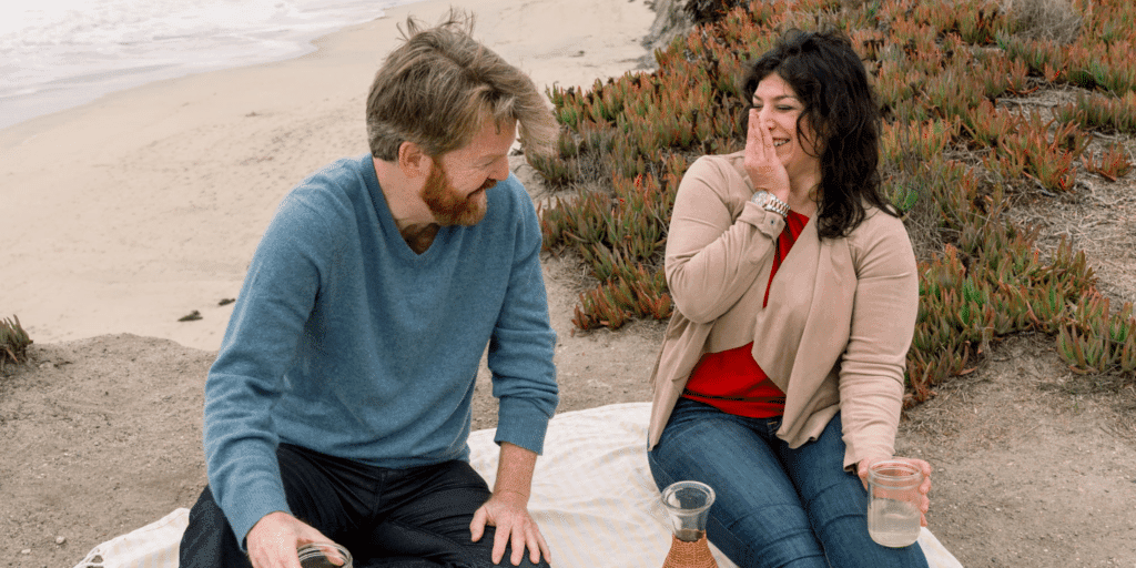East Bay date ideas. A man and woman laugh together while sipping drinks at a picnic on the beach.
