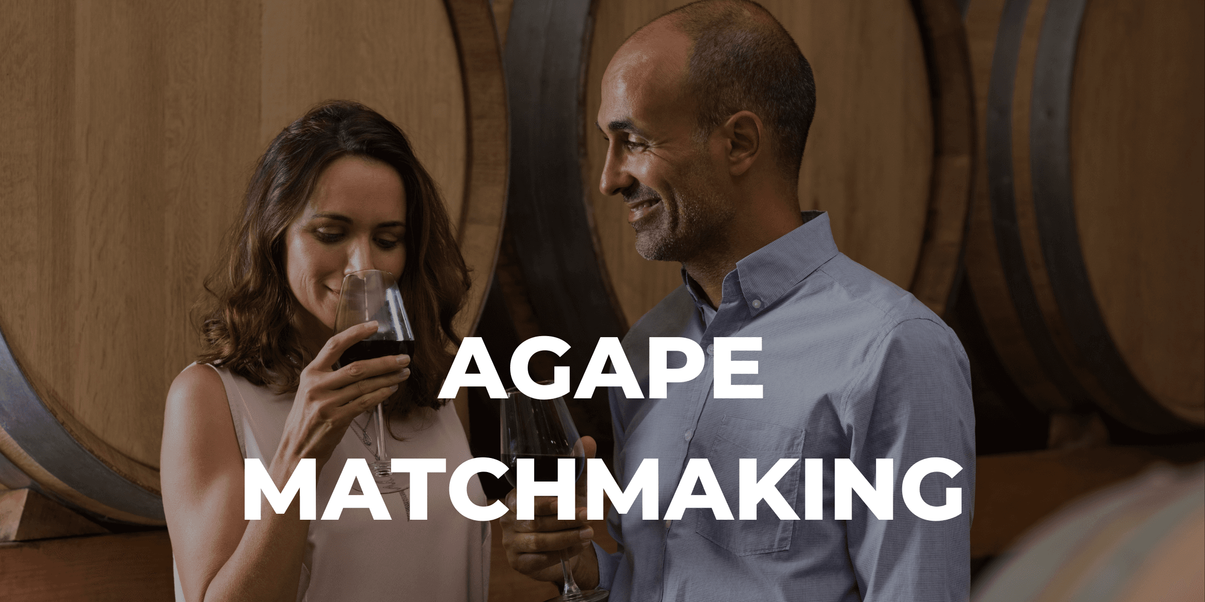 Image for Agape Matchmaking service showing a middle-aged man and woman holding glasses of red wine at a winery standing among wine barrels.