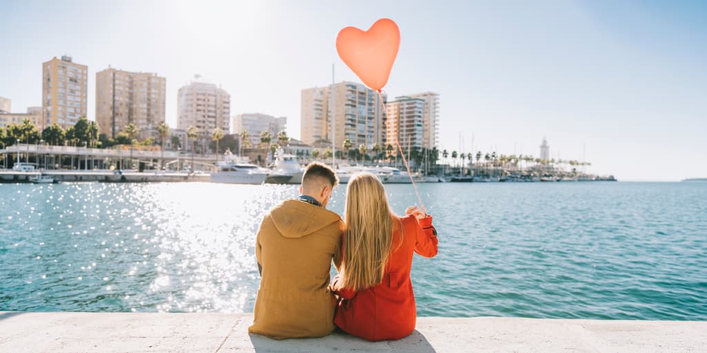A couple sits side by side on a sunny waterfront ledge holding a heart-shaped balloon, overlooking a marina and city skyline.
