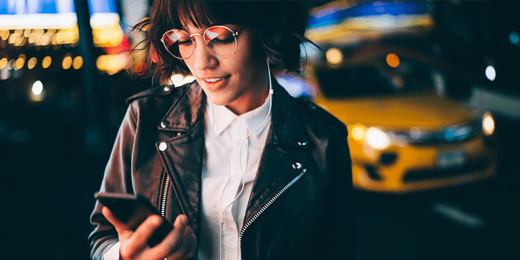 Woman in a city at night looking at her phone with a yellow taxi in the background, representing the fast-paced experience of using dating apps in New York City.