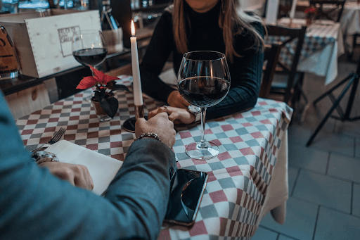 A heterosexual couple holds hands over a restaurant table with blue, red, and white checkerboard tablecloth, lit candlestick flower in a pot, and a glass of red wine. The man has his napkin and phone set on the table in front of him.