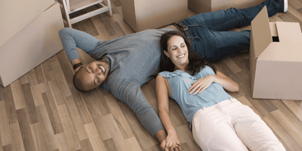 A man and a woman laying on the flower with moving boxes, holding hands and smiling.