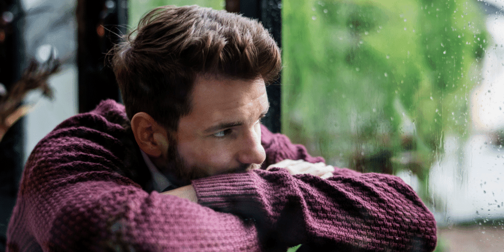 Is your man afraid to commit to your relationship? A man in a purple sweater appears sad as he looks out a window on a rainy day.